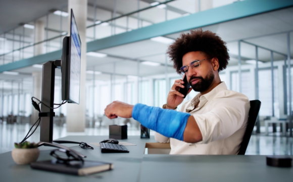 A man with an arm cast sits at a desk in a modern office, talking on the phone. He appears focused, surrounded by computer equipment and office supplies.